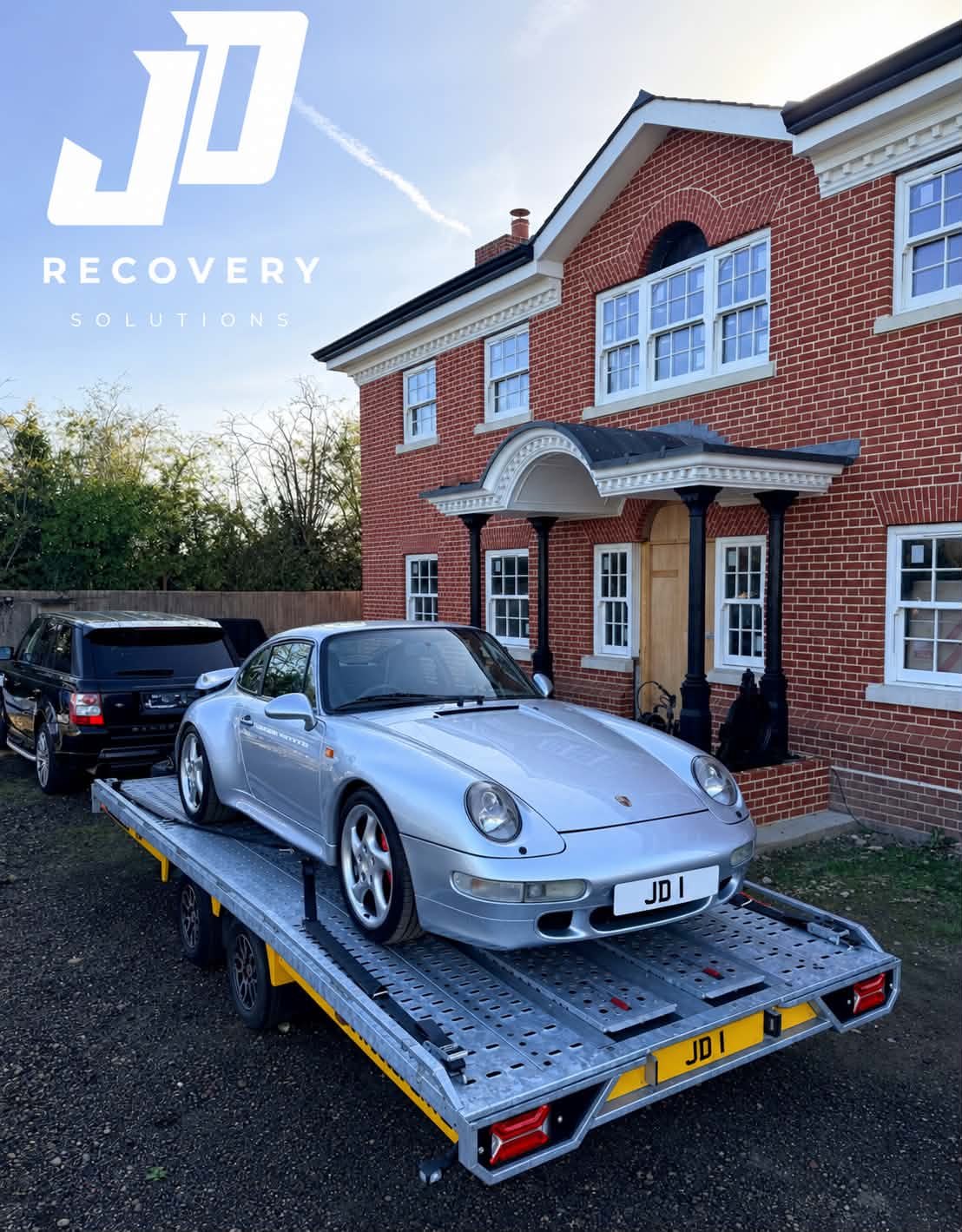 JD Recovery Solutions: silver Porsche 911 with JD 1 plates on a JD car transporter trailer outside a red-brick house